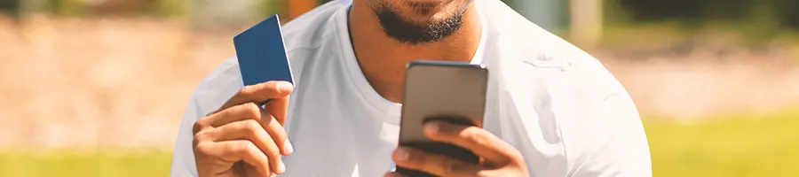 A person holding a credit card while looking at his phone outside in the park